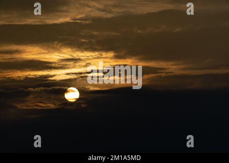 The setting winter sun is visible as a bright disk between dark scattered clouds Stock Photo