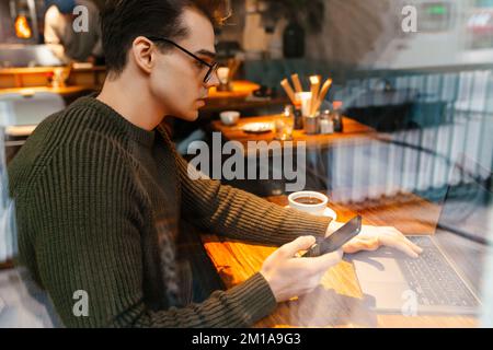 Young white man wearing eyeglasses using cellphone and laptop in cafe Stock Photo
