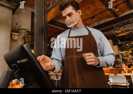 Young white waiter man wearing apron standing in front of computer and registering payment in cafe Stock Photo