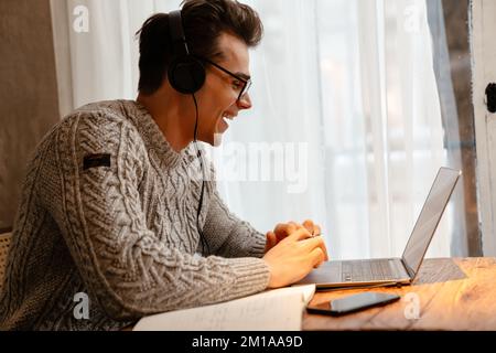 Young white man wearing eyeglasses smiling while using laptop and headphones in cafe Stock Photo