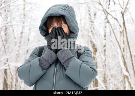 Frost weather in winter, woman in jacket standing in park during snow and covering her face by hands in black leather gloves Stock Photo