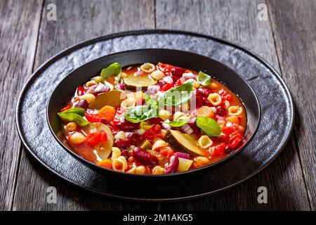 minestrone soup with red beans, zucchini, garbanzo beans, small pasta shells, cheese, tomato, carrots and basil in black bowl Stock Photo