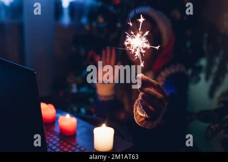 Online Christmas party. Celebrating New year from distance using laptop and internet. Woman burns sparkler and candles at home Stock Photo