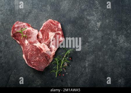 Raw beef steak. Marbled raw fresh Ribeye steak in heart shape with rosemary, salt and pepper on cutting board on dark concrete background. Raw beef st Stock Photo