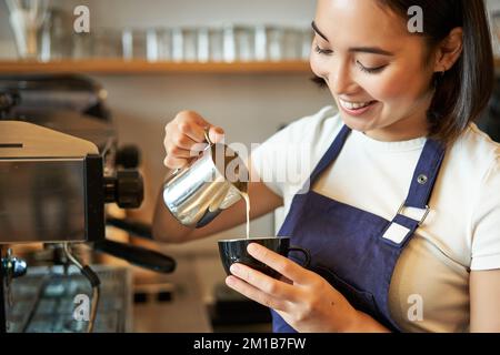 Smiling asian barista girl, making coffee, pouring steamed milk into cappuccino, doing latte art in cup, working in cafe Stock Photo