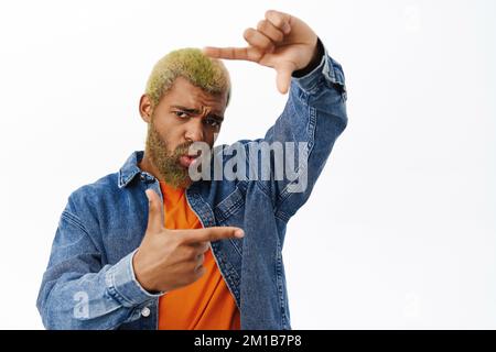 Snap. Stylish young man takes picture of you, looks through finger frames and smiles happy, searching for perfect angle, picturing smth, stands over Stock Photo