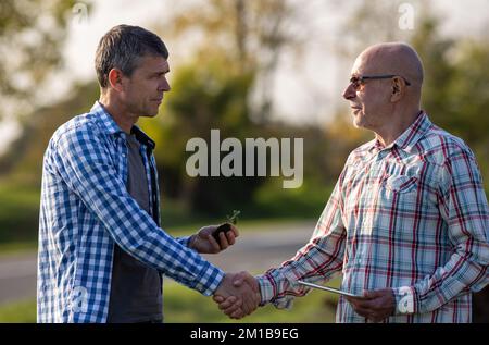 Two mature farmers shaking hands in field, holding sprout and checking crop growth Stock Photo