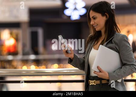 Side view of positive adult businesswoman with long dark hair in elegant suit smiling while messaging on smartphone  Stock Photo