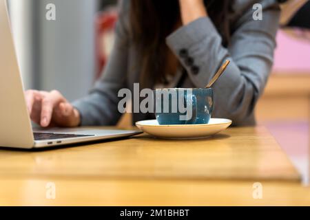 Crop anonymous female entrepreneur with long dark hair in gray suit working on laptop while sitting at wooden table with cup of cappuccino Stock Photo