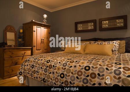 King size bed with flowery motifs bedspread in master bedroom with antique wood dresser and cabinet inside split-level style home. Stock Photo
