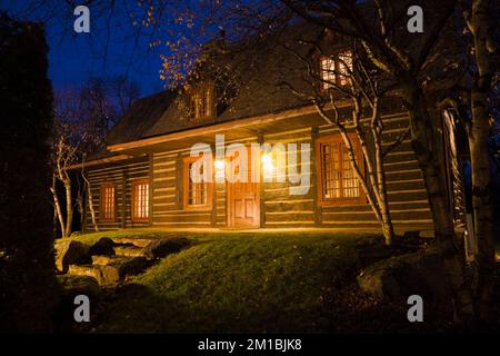 1975 built replica of old 1800s Canadiana cottage style log cabin illuminated at dusk. Stock Photo