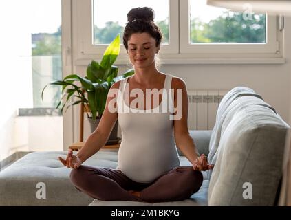 Pregnant woman sitting in lotus position and meditating on sofa in living room Stock Photo