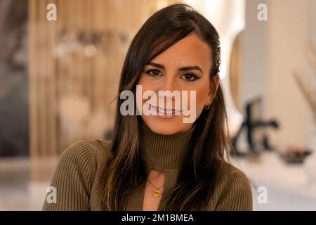 Optimistic adult woman in turtleneck with stylish makeup and long dark hair smiling and looking at camera on blurred background of beauty salon Stock Photo