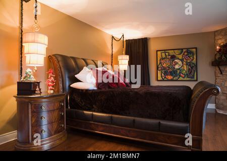 King size wood and leather sleigh bed and bedside table in master bedroom with tan yellow painted walls on upstairs floor inside luxurious home. Stock Photo
