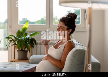 Pretty pregnant woman sitting on sofa, resting and drinking water on sofa in living room Stock Photo