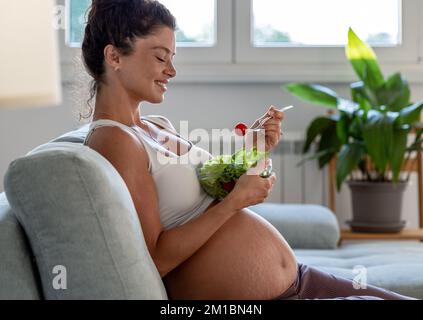Pretty young woman eating salad on sofa at home. Healthy lifestyle during maternity period Stock Photo