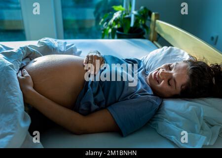 Pretty pregnant woman lying in bed, waking up, looking at belly and feeling happy Stock Photo