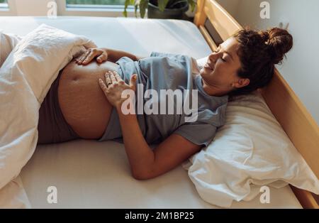 Pretty pregnant woman lying in bed, waking up, looking at belly and feeling happy Stock Photo