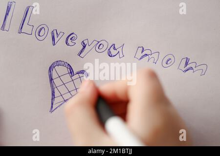 Mothers day.I love you mom inscription on a white sheet and a heart and a childs hand with a pen. Holiday of all mothers.moms day concept.  Stock Photo