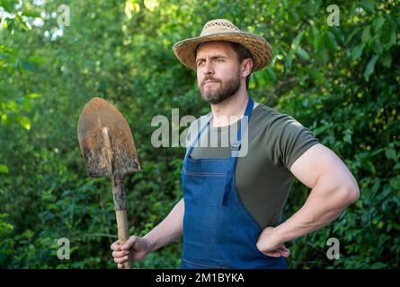 Thoughtful farmer man in farmers hat and gardening apron holding garden shovel natural outdoors. Stock Photo