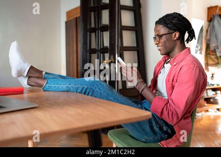 Smiling African guy freelancer using smartphone during working hours while working from home Stock Photo