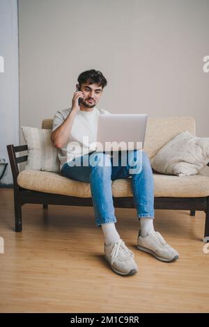 Joyful man freelancer having phone conversation while working at laptop, looking at laptop screen. Businessman talking with clients on mobile phone Stock Photo
