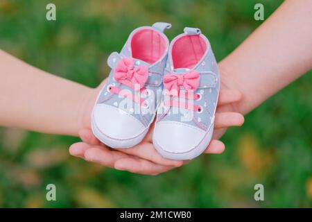 Close-up of blue and pink baby girl shoes joined by expecting parents' hands. Sneakers have bows, tiny hearts. Green background. Pregnancy announcement. Stock Photo