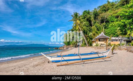 A traditional Filipino wooden outrigger boat called a banca sits on a beautiful tropical island sandy beach near resort buildings on Mindoro Island. Stock Photo