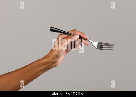 Crop anonymous African American female with silver metal dinner fork on gray background Stock Photo