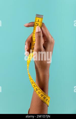 Crop anonymous African American female demonstrating yellow measuring tape wrapped around arm on turquoise background Stock Photo