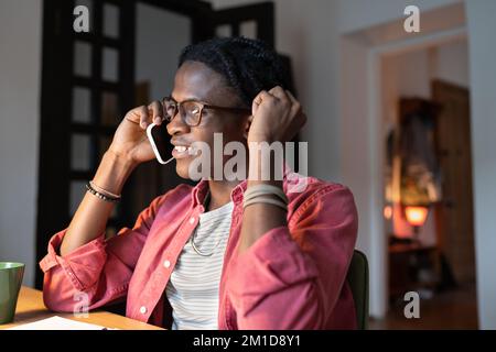 Smiling African guy college student talking on phone while studying at home, taking break from study Stock Photo