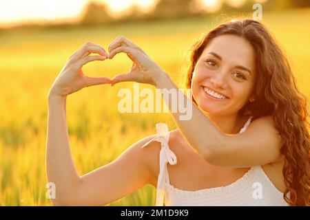 Happy woman making heart shape with hands at sunset in a wheat field Stock Photo