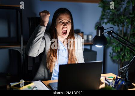 Young brunette woman working at the office at night angry and mad raising fist frustrated and furious while shouting with anger. rage and aggressive c Stock Photo