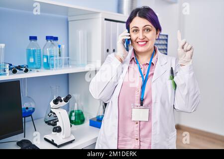 Plus size woman wit purple hair working at scientist laboratory speaking on g the phone smiling with an idea or question pointing finger with happy fa Stock Photo