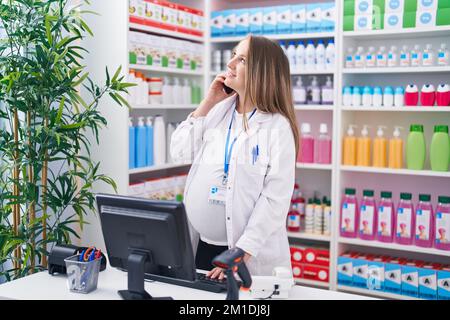 Young pregnant woman pharmacist talking on smartphone using computer at pharmacy Stock Photo