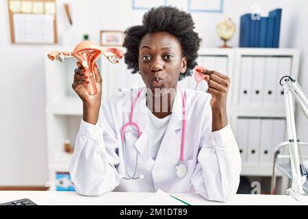 African doctor woman holding anatomical female genital organ and menstrual cup puffing cheeks with funny face. mouth inflated with air, catching air. Stock Photo