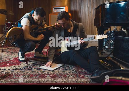 Two focused guitarists holding their guitars and correcting note sheet while sitting on the patterned carpet in their studio. High quality photo Stock Photo