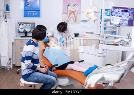 Kid patient in dentistiry office getting treatment for teeth cavity sitting on dental chair wearing bib. Dentistry specialist during child cavity consultation in stomatology office using modern technology. Stock Photo