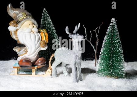 A toy shining silver deer carrying a sleigh with Santa Claus with gifts through the snowy forest among the Christmas trees at night. Christmas and New Stock Photo