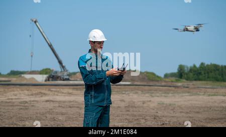 A man in a helmet and overalls controls a drone at a construction site. The builder carries out technical oversight. Stock Photo
