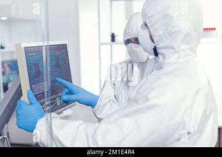 Biochemists in coverall typing on digital display the changes in vaccine composition in equipped laboratory. Doctors examining virus evolution using high tech researching diagnosis against covid19 Stock Photo