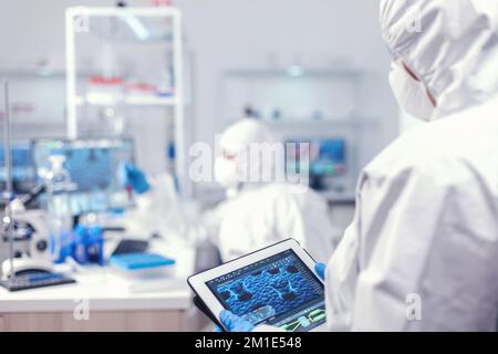 Doctor in ppe suit during coronavirus outbreak working in laboratory holding tablet pc. Team of scientists conducting vaccine development using high tech technology for researching treatment against covid19 Stock Photo