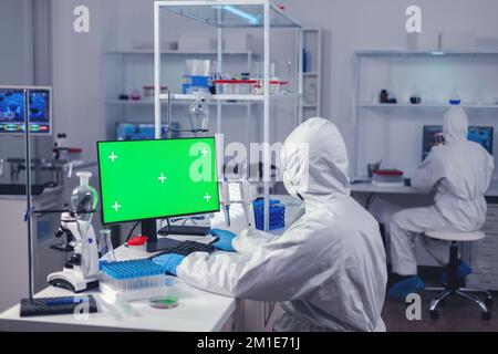 Doctor in protection suit against covid19 delevoping cure using computer with green screen dressed in ppe. Team of microbiologists doing vaccine research writing on device with chroma key, isolated, mockup display. Stock Photo