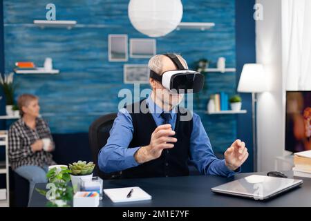 Senior man wearing virtual reality headset doing hand gesturing. Elderly man pensioner in living room sitting at desk with futuristic goggles for augmented cyberspace wife watching tv. Stock Photo