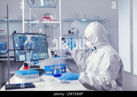 Concentrated scientist using micropipette with a test tube and coworker standing in the background. Chemist in modern laboratory doing research using dispenser during global epidemic with covid-19. Stock Photo