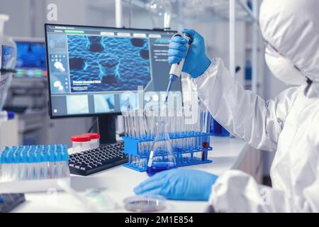 Scientist in coverall developing vaccine for coronavirus using automatic pipette. Chemist in modern laboratory doing research using dispenser during global epidemic with covid-19. Stock Photo