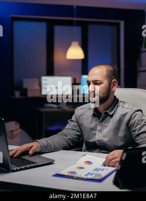 Frelancer working overtime in corporate workplace looking at laptop sitting at desk. Bearded entrepreneur doing overhourse at the office in the evening to finish a deadline. Stock Photo