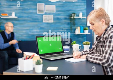 Confident senior woman using laptop Elderly woman working on computer with green screen and husband holding tv remote control. Stock Photo