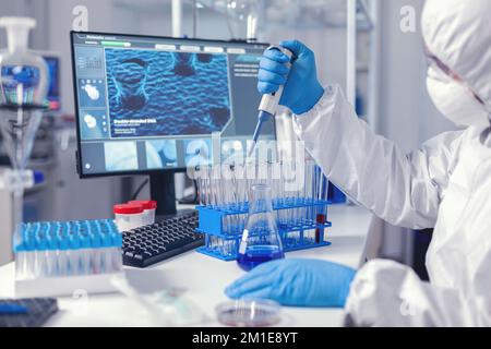 Laborant dropping sample in test tube using micropipette dressed in coverall. Chemist in modern laboratory doing research using dispenser during global epidemic with covid-19. Stock Photo