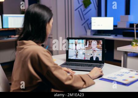 Entrepreneur talking in the course of online meeting with colleagues doing overtime. Woman working on finance during a video conference with coworkers at night hours in the office. Stock Photo